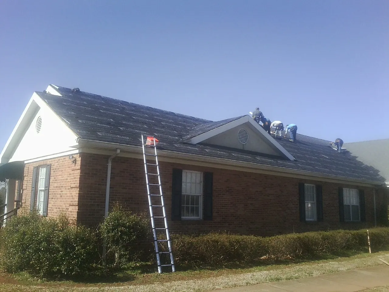 Crew tearing shingles off a brick ranch roof with a ladder set — Metal Roof Repair work by our crew in Unionville