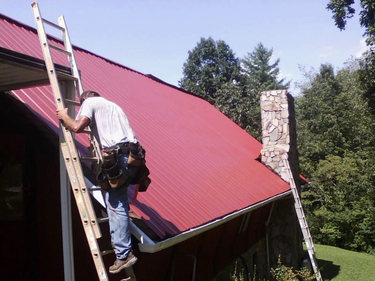 Roofer on a ladder beside a red metal roof and stone chimney — Soffit & Fascia Repair by Roofing Brothers in Unionville